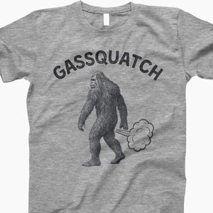 Puede incluir: Una camiseta gris jaspeado con un gr&aacute;fico de una criatura parecida a Pie Grande emitiendo una nube de gas. El texto "Gassquatch" est&aacute; arqueado sobre la ilustraci&oacute;n en negro. La camiseta tiene cuello redondo y mangas cortas.