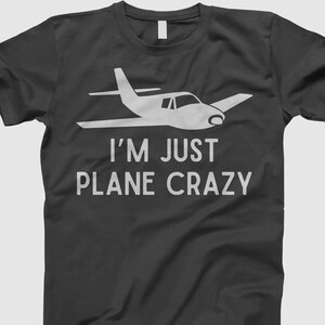 May include: Dark gray t-shirt with a white graphic of a small airplane and the text "I'M JUST PLANE CRAZY". The design features a simple, clean illustration of an aircraft, perfect for aviation enthusiasts.