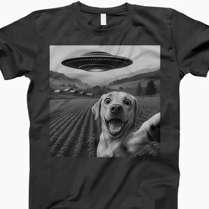 May include: Black t-shirt featuring a grayscale image of a dog taking a selfie in a field with a UFO hovering above. The dog has a wide, excited grin. The background includes a rural landscape with fields and buildings.
