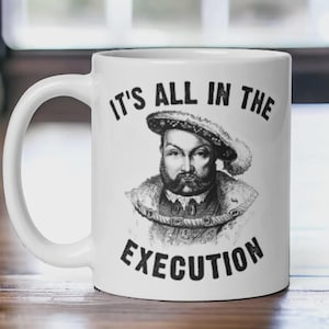May include: White ceramic mug featuring a black and white portrait of a historical figure with the text "IT'S ALL IN THE EXECUTION". The mug has a curved handle and is set against a blurred background.