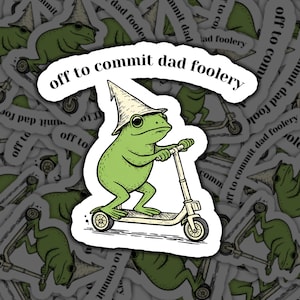 May include: A sticker featuring a green frog wearing a wizard hat and riding a scooter. The text "off to commit dad foolery" is arched above the frog. The design has a whimsical, cartoonish style.