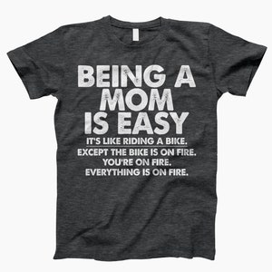 May include: Dark heather grey t-shirt with a white graphic that reads "Being a Mom is Easy. It's like riding a bike. Except the bike is on fire. You're on fire. Everything is on fire."