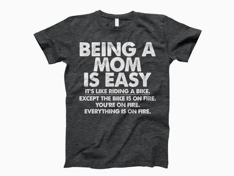 May include: Dark heather grey t-shirt with a white graphic that reads "Being a Mom is Easy. It's like riding a bike. Except the bike is on fire. You're on fire. Everything is on fire."