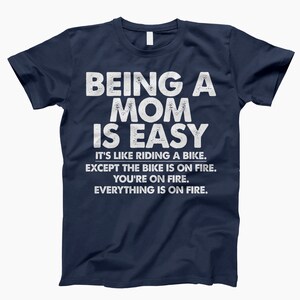 May include: Navy blue t-shirt with white text that reads "Being a Mom is Easy. It's like riding a bike. Except the bike is on fire. You're on fire. Everything is on fire."