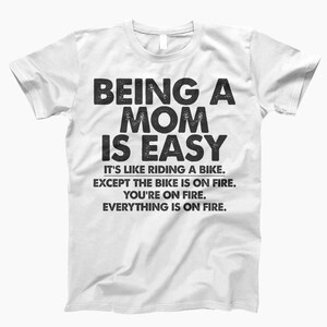 May include: White t-shirt with black text that reads "Being a Mom is Easy. It's like riding a bike, except the bike is on fire. You're on fire. Everything is on fire."