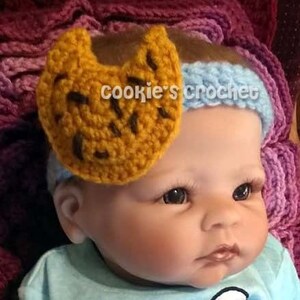 May include: A baby wearing a light blue headband with a crocheted brown and yellow cookie attached to it.