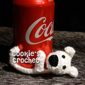 May include: White crocheted dog toy with black eyes and nose, laying on a wooden surface next to a red can of Coca-Cola. The can has the text "Coca-Cola" and "Original" on it. The toy has the text "Cookie's Crochet" on it.