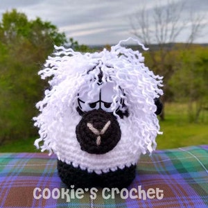May include: A white crocheted sheep with black eyes and a black nose. The sheep has a fluffy white mane and is sitting on a tartan surface.