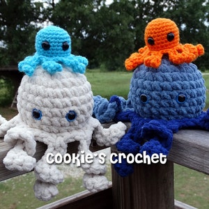 May include: Two crocheted octopus plushies, one white with a blue octopus on top and one blue with an orange octopus on top. The plushies are sitting on a wooden railing. "cookie's crochet" is written on the railing.