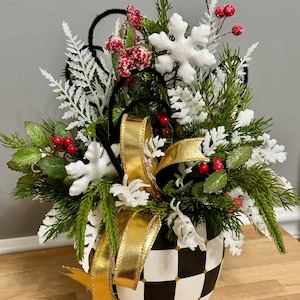 May include: A festive arrangement in a black and white checkered vase. The arrangement features green pine branches, white snowflake accents, red berries, and a gold ribbon. A black handle arches over the top of the arrangement.