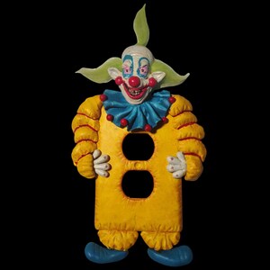 May include: A yellow clown-shaped light switch cover with a smiling face, red nose, and blue ruffled collar. The clown has green hair and blue shoes. The cover has two circular openings for outlets.