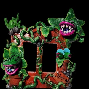 May include: A handmade light switch cover featuring a brick wall with three carnivorous plants. The plants have green leaves, pink mouths with teeth, and are in brown pots. The switch cover has a sign that says "Feed Me".