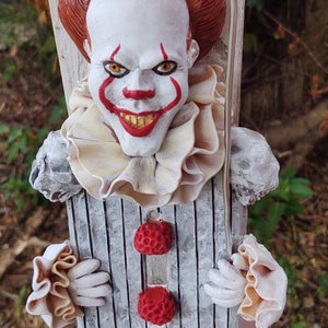 May include: A detailed, hand-painted clown figure with a menacing expression, red hair, and a red balloon. The clown has a white face with red accents, a ruffled collar, and a striped torso. Two red pom-poms adorn the front.
