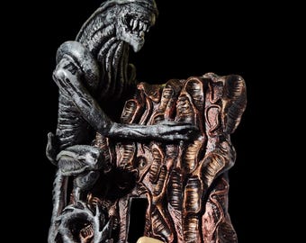 Può includere: Una statuetta aliena argentata e nera posata su una base testurizzata di colore rame. L'alieno si tiene alla base con le mani. Un uovo alieno rosa e verde si trova nella parte inferiore della base.