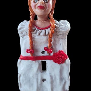 May include: A white porcelain light switch cover with a red ribbon and a red rose. The cover is decorated with a doll with red hair, a white dress, and a creepy expression.