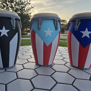 May include: Three conga drums featuring the Puerto Rican flag design. The left drum is black, white, and gray. The middle drum is light blue, red, and white. The right drum is dark blue, red, and white. Each drum has a white star and gray hardware.