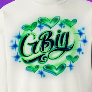 May include: White sweatshirt with a green and blue airbrushed design featuring the text "G Big" surrounded by hearts and stars.