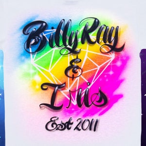 May include: White t-shirt with an airbrushed design of a rainbow diamond with the text "Billy Ray & Tris Est 2011" in black lettering. The design is surrounded by a pink, blue, and yellow watercolor effect.