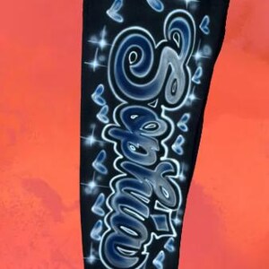 Puede incluir: Pantalones deportivos negros con un dise&ntilde;o personalizado pintado con aer&oacute;grafo. El dise&ntilde;o presenta el nombre "Samie" en una fuente estilizada, acentuado con mariposas azules y destellos brillantes. Los pantalones tienen cintura el&aacute;stica.