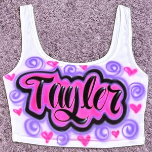 Może przedstawiać: Biały top z napisem "Taylor" w stylu graffiti. Nazwa jest czarna z r&oacute;żowym obrysem, na r&oacute;żowo-fioletowym tle chmur z sercami i wirami.