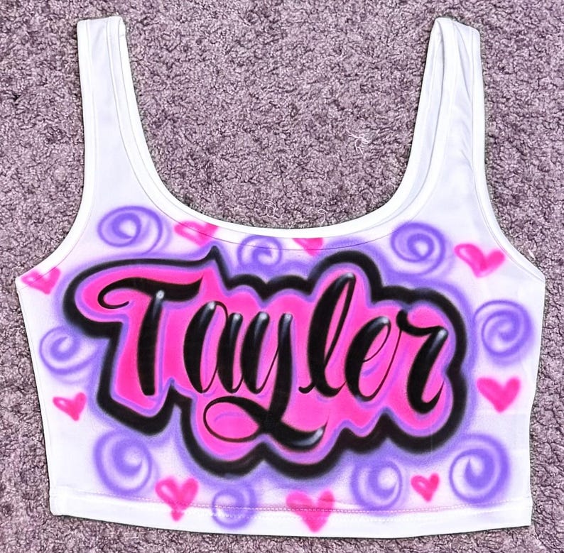 Może przedstawiać: Biały top z napisem "Taylor" w stylu graffiti. Nazwa jest czarna z r&oacute;żowym obrysem, na r&oacute;żowo-fioletowym tle chmur z sercami i wirami.