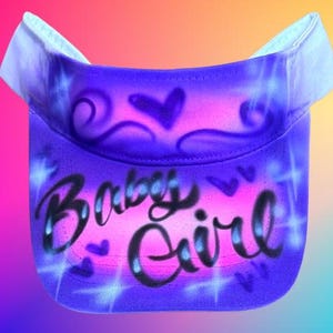 May include: A purple visor featuring the words "Baby Girl" in black script. The visor has a gradient design with pink and blue hues, hearts, and starburst accents.