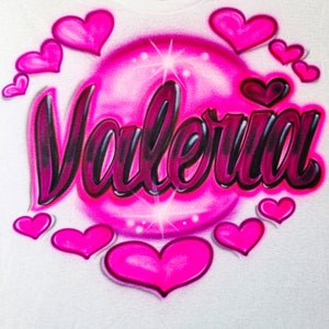 May include: A white t-shirt featuring a pink bubble design with the name "Valeria" written in black and pink lettering. The bubble is surrounded by pink hearts.