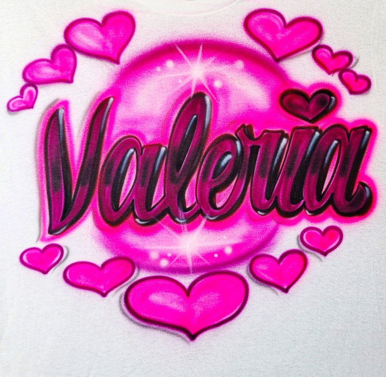May include: A white t-shirt featuring a pink bubble design with the name "Valeria" written in black and pink lettering. The bubble is surrounded by pink hearts.