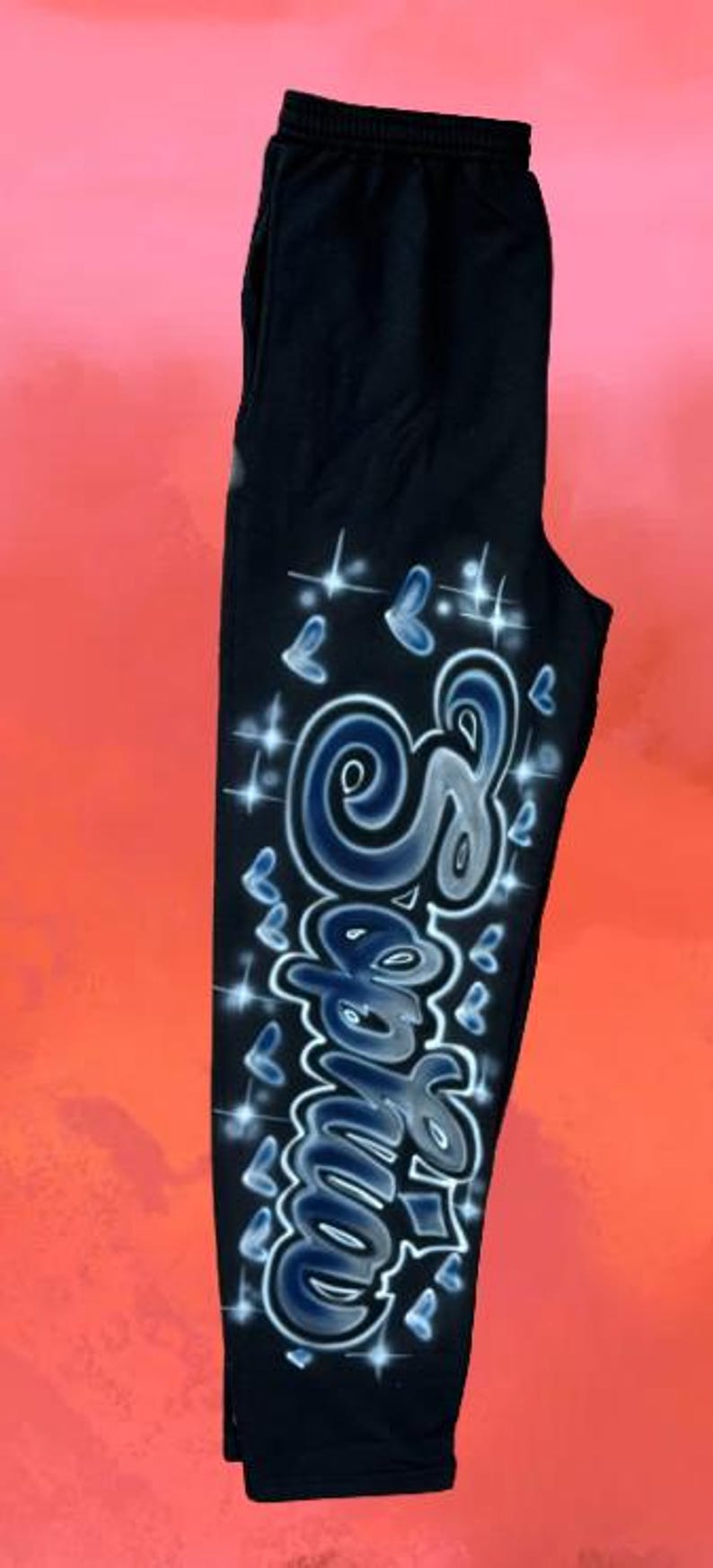 Puede incluir: Pantalones deportivos negros con un dise&ntilde;o personalizado pintado con aer&oacute;grafo. El dise&ntilde;o presenta el nombre "Samie" en una fuente estilizada, acentuado con mariposas azules y destellos brillantes. Los pantalones tienen cintura el&aacute;stica.