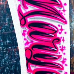 Puede incluir: Un panel de tela blanco con la palabra "Love" escrita en letras grandes, estilizadas, negras y rosas. Las letras est&aacute;n delineadas en negro y rellenas de rosa. Hay peque&ntilde;as estrellas rosas y blancas dispersas alrededor de las letras.