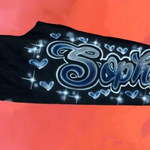 Puede incluir: Pantalones deportivos negros con el nombre "Sophia" en una fuente estilizada, aerografiada. El nombre est&aacute; delineado en blanco y sombreado con azul, rodeado de corazones azules y estrellas. Los pantalones se muestran sobre un fondo degradado rojo y rosa.