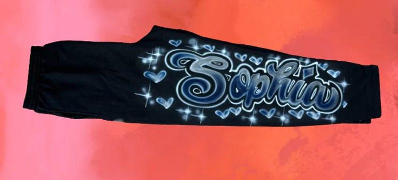 Puede incluir: Pantalones deportivos negros con el nombre "Sophia" en una fuente estilizada, aerografiada. El nombre est&aacute; delineado en blanco y sombreado con azul, rodeado de corazones azules y estrellas. Los pantalones se muestran sobre un fondo degradado rojo y rosa.