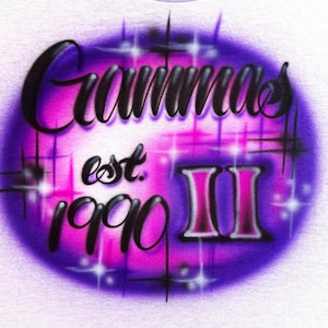Puede incluir: Camiseta blanca con un diseño aerografiado. El diseño presenta la palabra "Grammas" en escritura negra, "est. 1990" y el número romano "II" en un óvalo rosa y morado con detalles de estrellas.