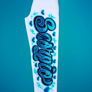 Puede incluir: Pantalones de ch&aacute;ndal blancos con un dise&ntilde;o gr&aacute;fico azul y negro de la palabra "Sophia" en la pierna derecha. El dise&ntilde;o incluye corazones y estrellas.