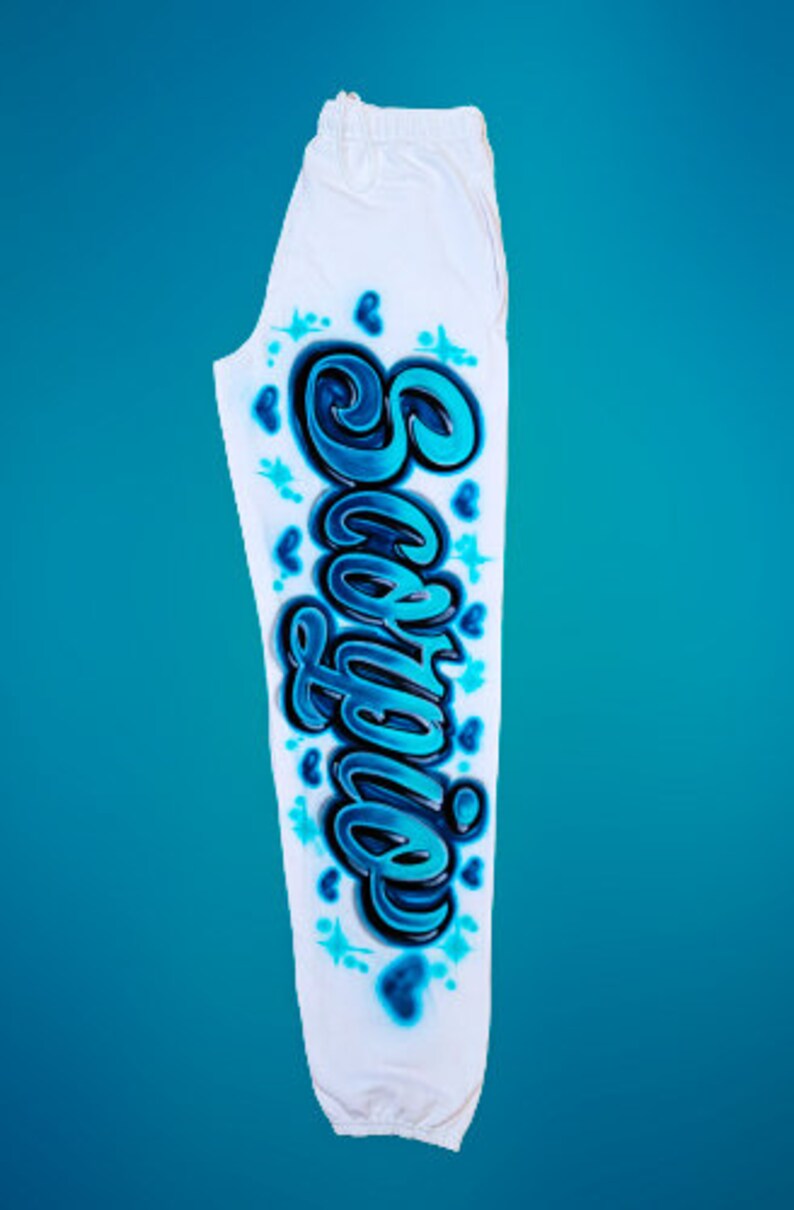 Puede incluir: Pantalones de ch&aacute;ndal blancos con un dise&ntilde;o gr&aacute;fico azul y negro de la palabra "Sophia" en la pierna derecha. El dise&ntilde;o incluye corazones y estrellas.