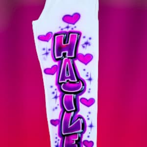 Puede incluir: Pantalones de ch&aacute;ndal blancos con un dise&ntilde;o de graffiti rosa y morado del nombre "HAILEY" con corazones y estrellas.