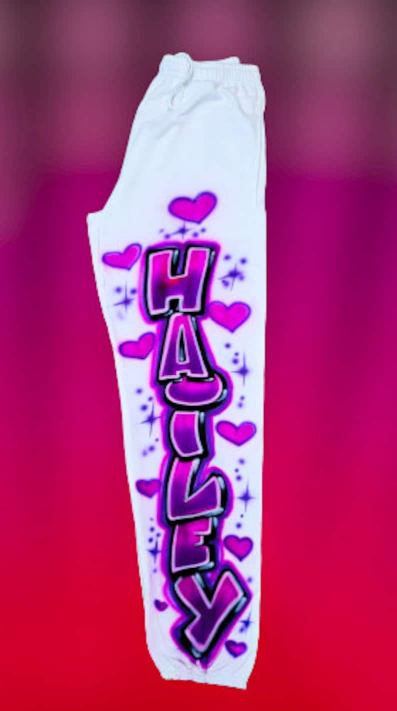 Puede incluir: Pantalones de ch&aacute;ndal blancos con un dise&ntilde;o de graffiti rosa y morado del nombre "HAILEY" con corazones y estrellas.