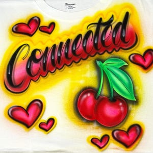以下が含まれることがあります： 白いTシャツにエアブラシのデザイン。デザインには、筆記体の「Connected」という単語、赤いさくらんぼ、赤いハートが含まれています。背景は黄色とオレンジ色です。
