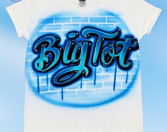 May include: White t-shirt with a blue and black airbrushed design of the words "Big Tot" on a brick wall background.