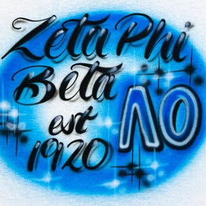 Op de afbeelding: Een airbrush-ontwerp op een witte achtergrond met de woorden "Zeta Phi Beta" in zwart script, met "est 1920" eronder. De tekst staat tegen een blauwe ovaal met sprankelende accenten en de letters "ΛO" in blauw.