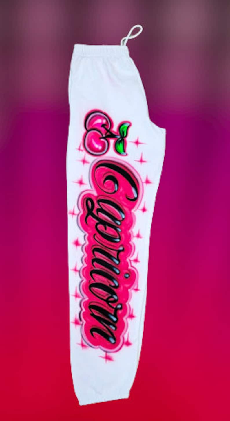 Puede incluir: Pantalones de ch&aacute;ndal blancos con una cereza rosa y la palabra "Capricornio" escrita en letras negras y rosas.
