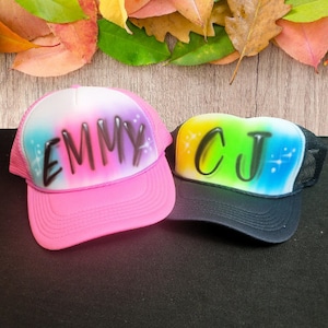 Puede incluir: Dos gorras de camionero coloridas con paneles traseros de malla. La gorra rosa tiene un diseño degradado arcoíris con el nombre "EMMY" en letras negras. La gorra negra tiene un diseño degradado arcoíris con las iniciales "CJ" en letras negras.