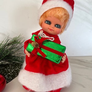 May include: A vintage Christmas doll wearing a red and white Santa suit and holding a green gift box with a red ribbon.