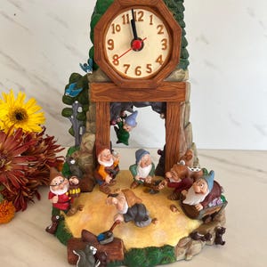 May include: A decorative clock featuring a scene with the seven dwarfs. The clock has a wooden-style frame with a cream-colored clock face. The dwarfs are depicted in various poses, with a forest backdrop.