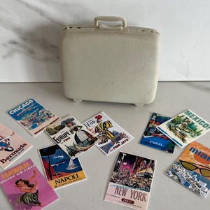 May include: A beige miniature suitcase with a handle, surrounded by travel-themed postcards. The postcards feature destinations like Chicago, Hawaii, and New York, with vintage airline logos and illustrations. The overall theme is travel and nostalgia.