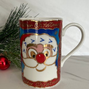 May include: A white ceramic mug with a red, blue, and gold illustration of Santa Claus's face. The mug has the words "Father Christmas" written in gold around the rim.