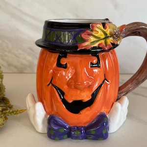 May include: Orange ceramic pumpkin mug with a smiling face, black eyes and mouth, and a purple bow tie. The mug has a black hat with green and purple accents and a brown handle. The mug is decorated with a fall leaf.