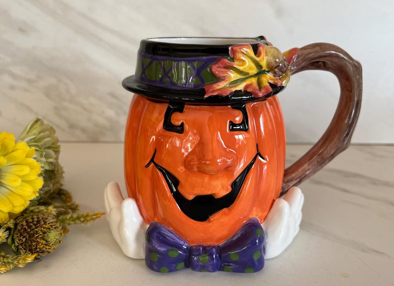May include: Orange ceramic pumpkin mug with a smiling face, black eyes and mouth, and a purple bow tie. The mug has a black hat with green and purple accents and a brown handle. The mug is decorated with a fall leaf.