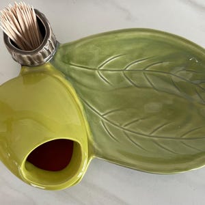 May include: A ceramic serving dish in the shape of a leaf, with a light green glaze. It has a small, round container for toothpicks and a larger compartment. The dish is approximately 12 inches long.