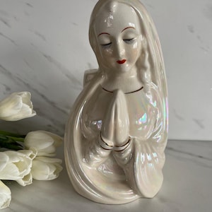 May include: A ceramic figurine of a woman with closed eyes and clasped hands. The figure is off-white with iridescent highlights, red lips, and gold accents. The woman is wearing a head covering. The figurine is a religious item.